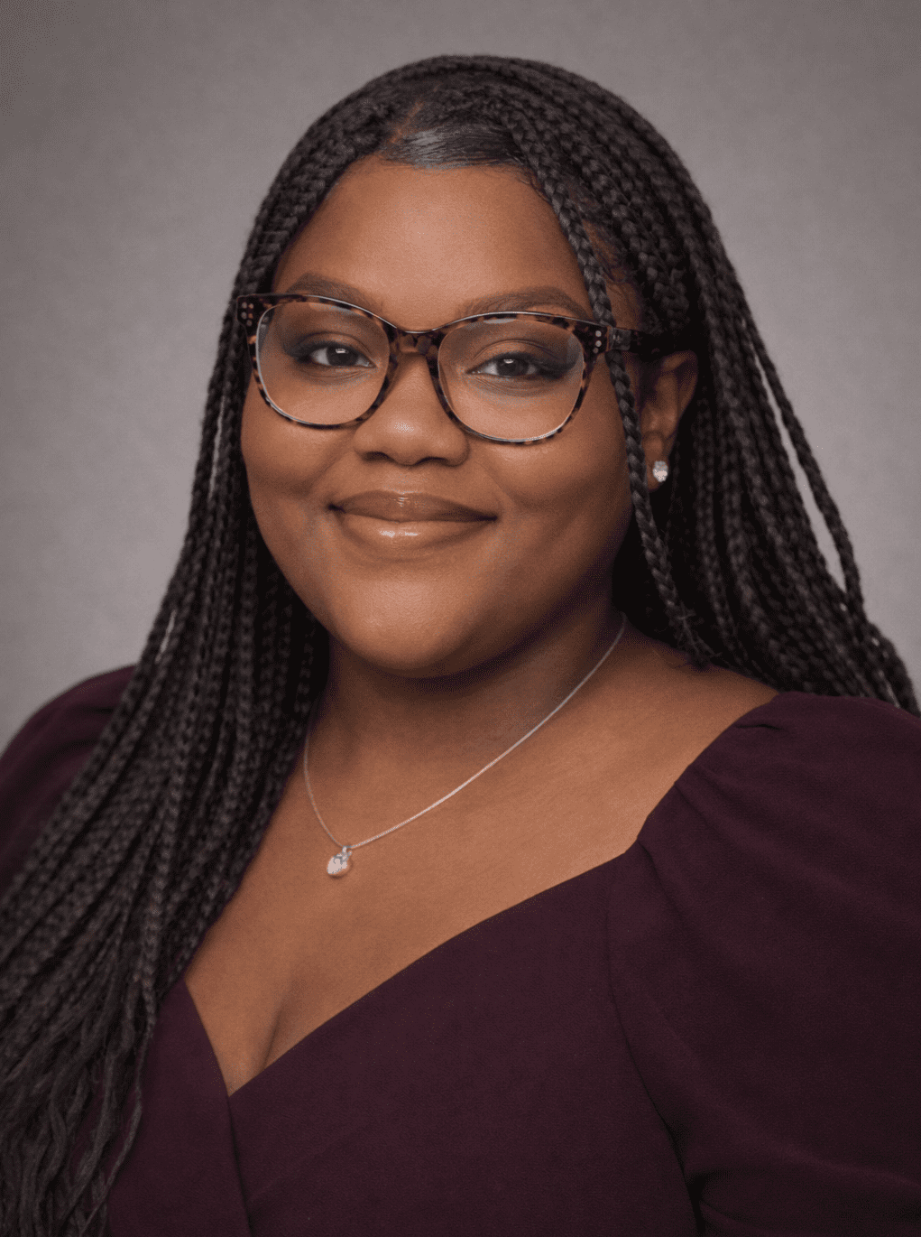A smiling woman with braided hair and glasses against a neutral background.